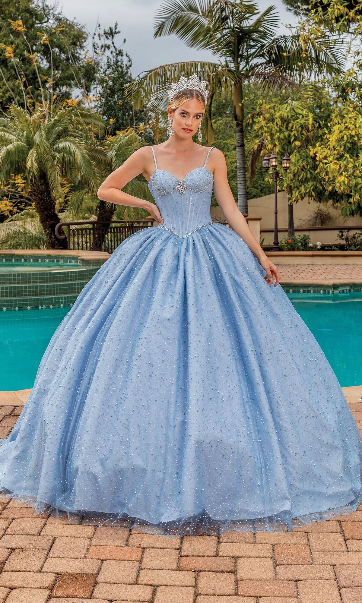 A woman stands by a pool in the Quinceanera Dress 1891 By Dancing Queen, featuring a fitted bodice, thin straps, and sparkling accents, accessorized with a tiara. Palm trees and lush greenery complete the scene.